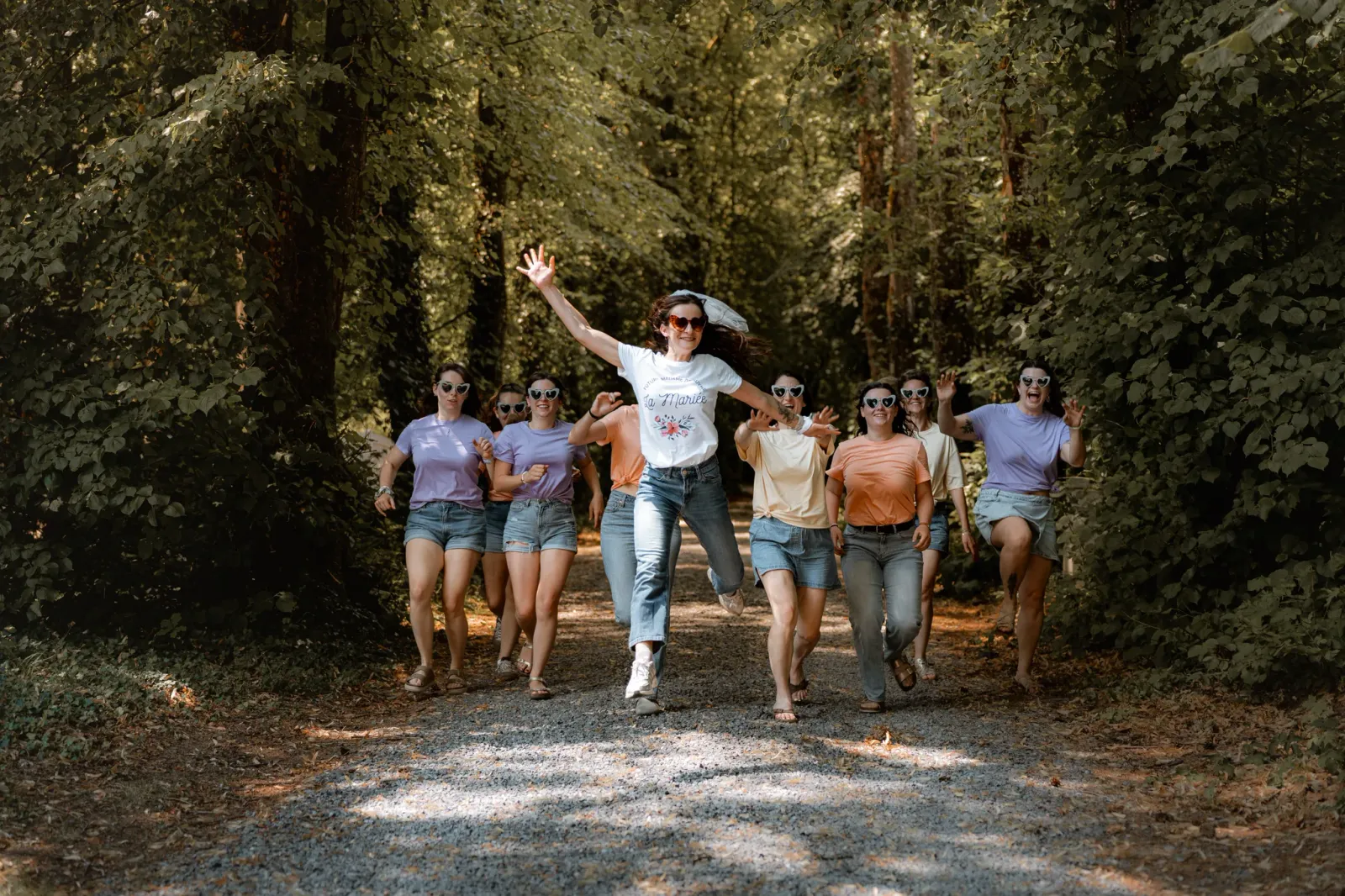 La future mariée saute de joie, bras levés, entourée de ses amies sur un chemin de forêt